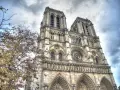 Notre Dame i Paris, ett mästerverk av romansk arkitektur, reser sig mot en dramatisk himmel. Trädgrenar med höstlöv ramar in den storslagna fasaden.