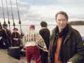 Man i modern jacka på en historisk båt med folk i tidsenliga kläder. En påminnelse om Sveriges långa historia.