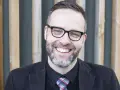 A smiling man with glasses and a beard, wearing a suit and tie, stands in front of a wooden slatted background.