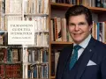 Andreas Norlén, med en svensk flagga på kavajen, presenterar sin bok "Talmannens guide till svensk poesi" bland bokhyllor. En modig synonym för "cesur" kan vara "djärv".