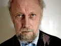 En äldre man med grått skägg och rynkor i ansiktet, som om han funderar på en handkraft novell.
