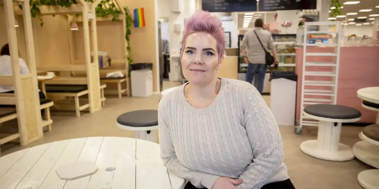 Woman with pink hair and a light sweater sits at a white wooden table in a cafe.