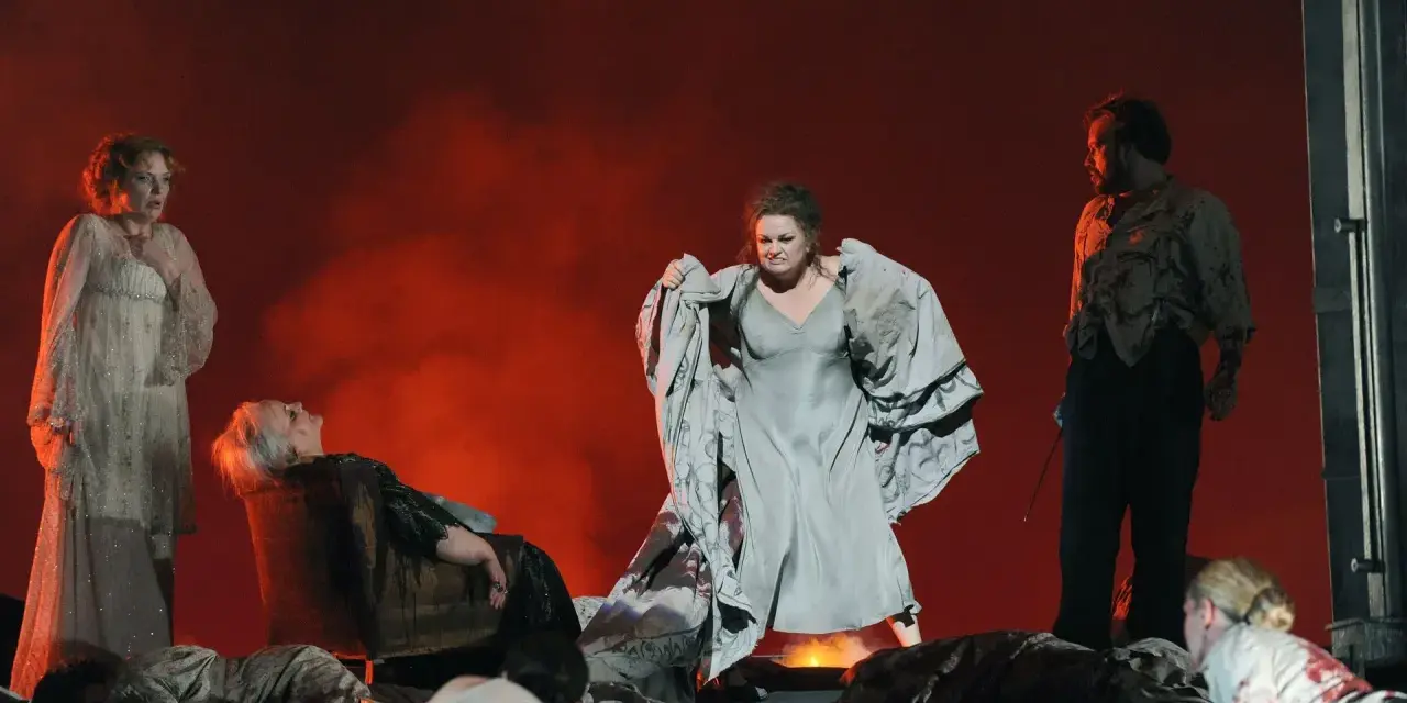 Scen från en grekisk teater med skådespelare i dramatiska poser. En kvinna i ljus klänning, en annan i grå klänning med tyg i famnen, och en man i mörk kostym.