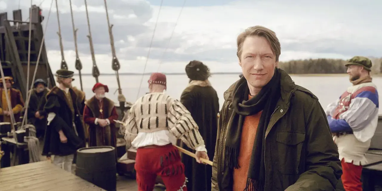 Man i modern jacka på en historisk båt med folk i tidsenliga kläder. En påminnelse om Sveriges långa historia.