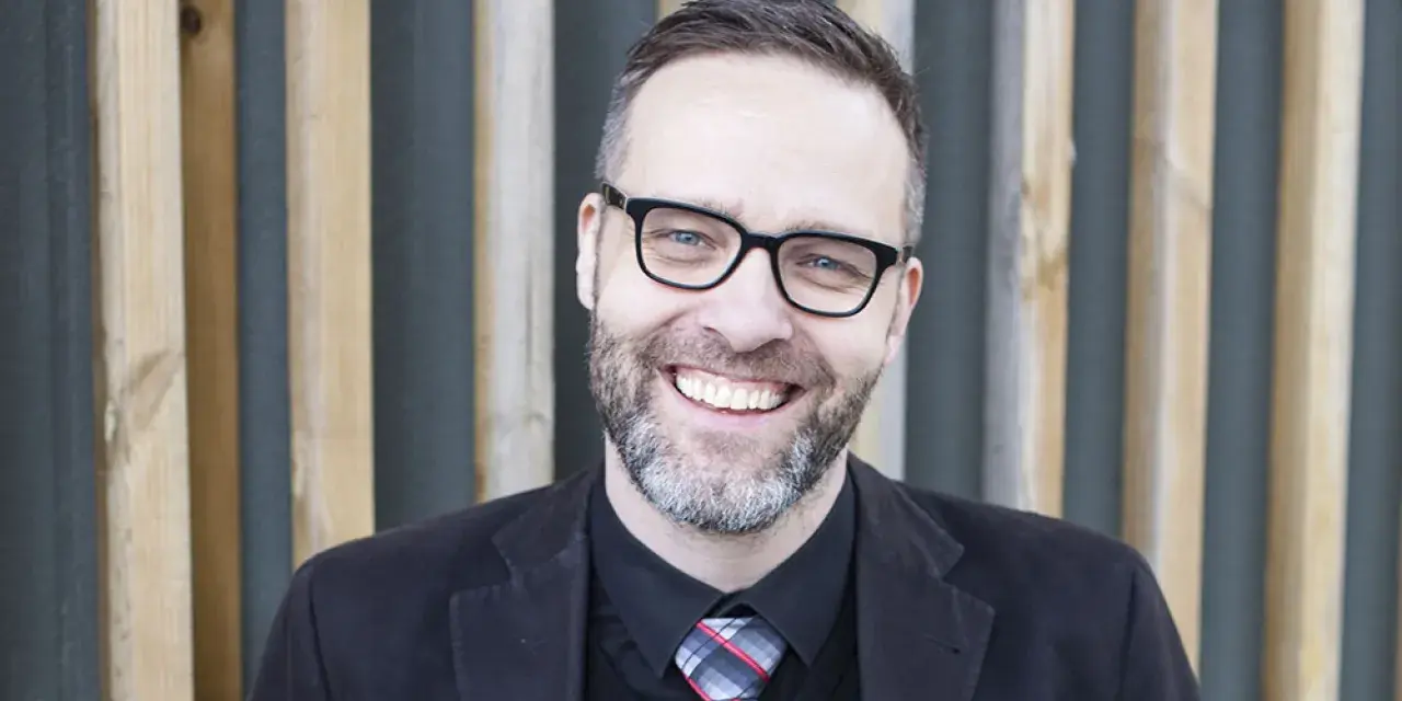 A smiling man with glasses and a beard, wearing a suit and tie, stands in front of a wooden slatted background.