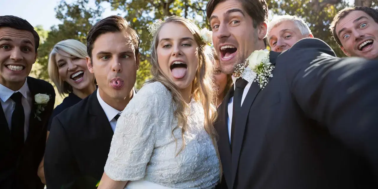 A wedding party takes a silly selfie outdoors, with the bride and groom sticking out their tongues.