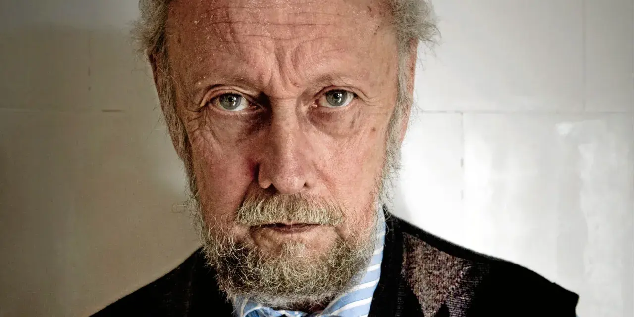 En äldre man med grått skägg och rynkor i ansiktet, som om han funderar på en handkraft novell.