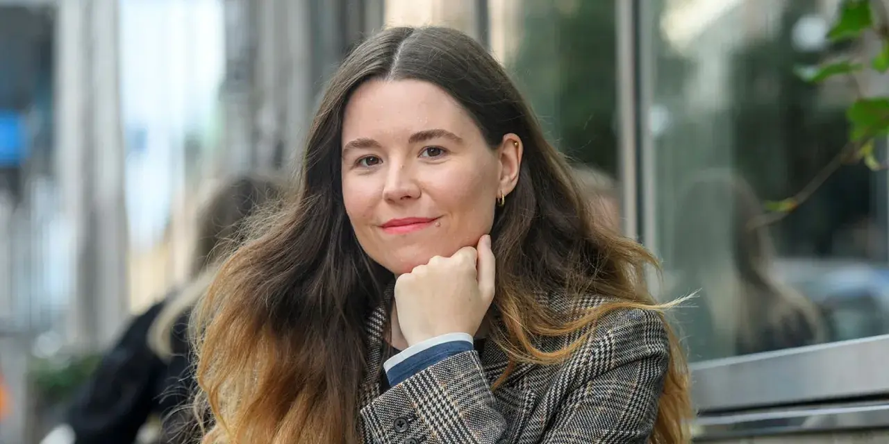 Johanna Hedman, med sitt långa, vågiga hår och en rutig kavaj, ler mot betraktaren.