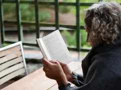 En person med lockigt hår läser en bok utomhus. Texten på sidan verkar vara en svensk dikt.