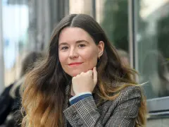 Johanna Hedman, med sitt långa, vågiga hår och en rutig kavaj, ler mot betraktaren.