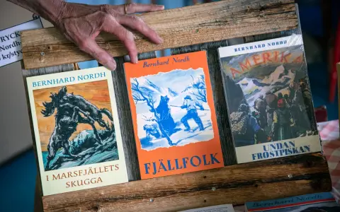 En hand vilar på tre böcker av Bernhard Nordh: "I Marsfjällets skugga", "Fjällfolk" och "Amerika – Undan frostpiskan".