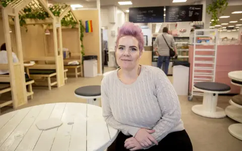 Woman with pink hair and a light sweater sits at a white wooden table in a cafe.