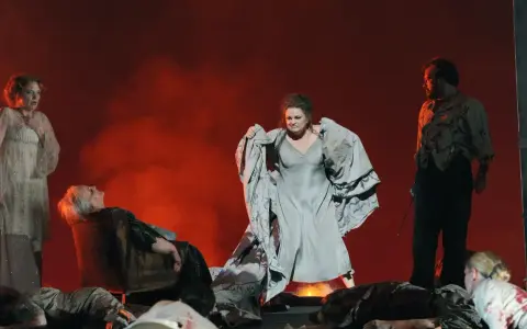 Scen från en grekisk teater med skådespelare i dramatiska poser. En kvinna i ljus klänning, en annan i grå klänning med tyg i famnen, och en man i mörk kostym.