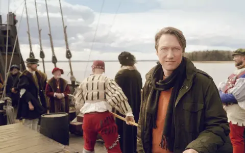 Man i modern jacka på en historisk båt med folk i tidsenliga kläder. En påminnelse om Sveriges långa historia.