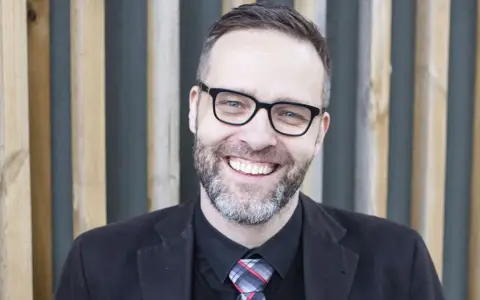 A smiling man with glasses and a beard, wearing a suit and tie, stands in front of a wooden slatted background.
