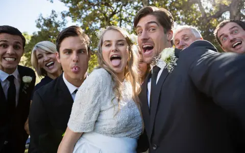 A wedding party takes a silly selfie outdoors, with the bride and groom sticking out their tongues.