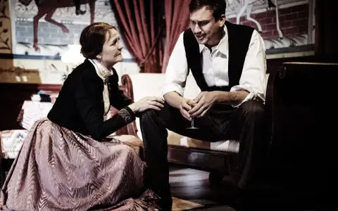 En kvinna i en stor klänning och en man i väst sitter nära varandra, en scen som påminner om ett ibsen drama.