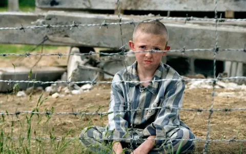 Pojken i randig pyjamas, Shmuel, sitter bakom taggtråd. Hans ansikte är smutsigt och hans blick är sorgsen.
