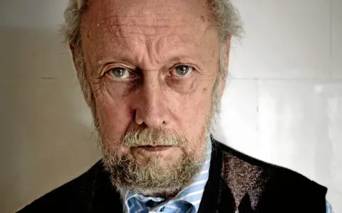 En äldre man med grått skägg och rynkor i ansiktet, som om han funderar på en handkraft novell.