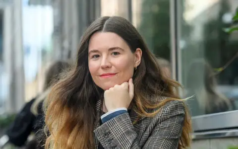 Johanna Hedman, med sitt långa, vågiga hår och en rutig kavaj, ler mot betraktaren.