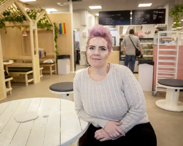 Woman with pink hair and a light sweater sits at a white wooden table in a cafe.