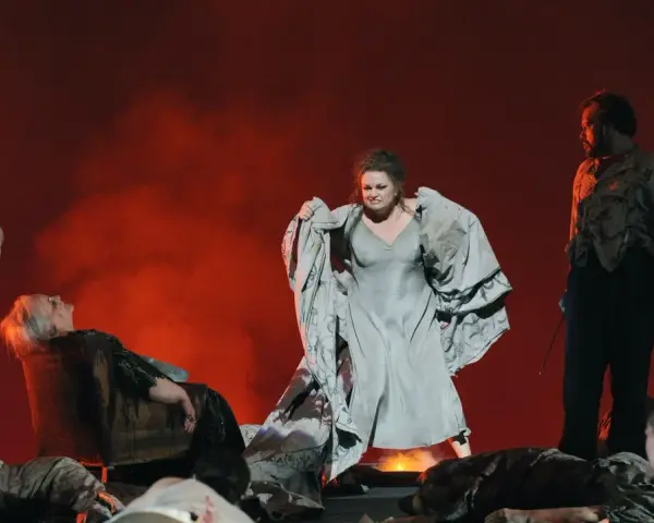 Scen från en grekisk teater med skådespelare i dramatiska poser. En kvinna i ljus klänning, en annan i grå klänning med tyg i famnen, och en man i mörk kostym.
