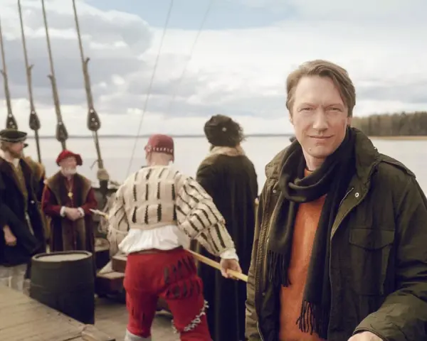 Man i modern jacka på en historisk båt med folk i tidsenliga kläder. En påminnelse om Sveriges långa historia.