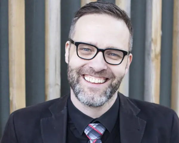 A smiling man with glasses and a beard, wearing a suit and tie, stands in front of a wooden slatted background.