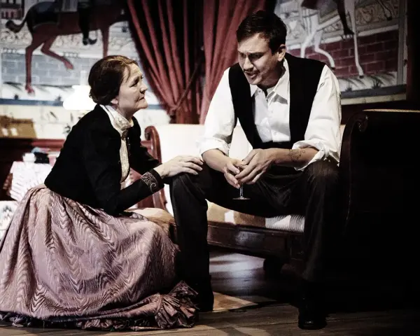 En kvinna i en stor klänning och en man i väst sitter nära varandra, en scen som påminner om ett ibsen drama.