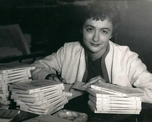 Françoise Sagan vid ett bord med högar av böcker, hon skriver på ett papper.