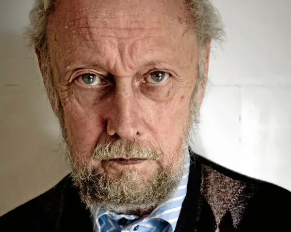 En äldre man med grått skägg och rynkor i ansiktet, som om han funderar på en handkraft novell.