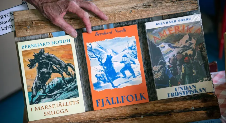 En hand vilar på tre böcker av Bernhard Nordh: "I Marsfjällets skugga", "Fjällfolk" och "Amerika – Undan frostpiskan".