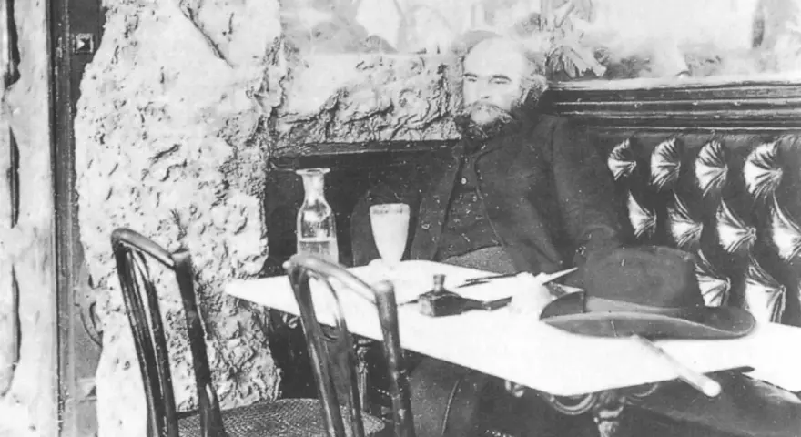 Paul Verlaine sitter vid ett bord med en hatt och en flaska.