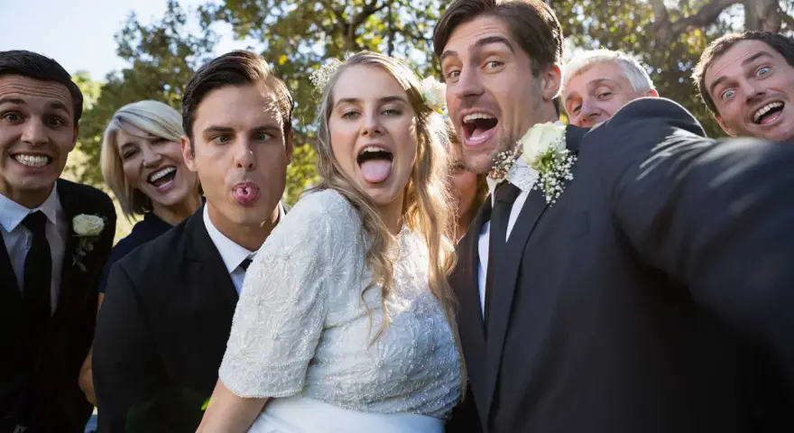 A wedding party takes a silly selfie outdoors, with the bride and groom sticking out their tongues.