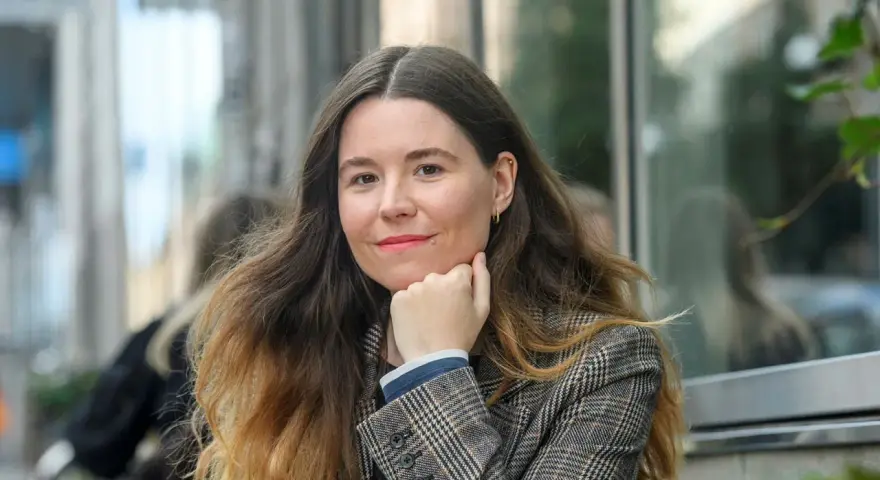 Johanna Hedman, med sitt långa, vågiga hår och en rutig kavaj, ler mot betraktaren.