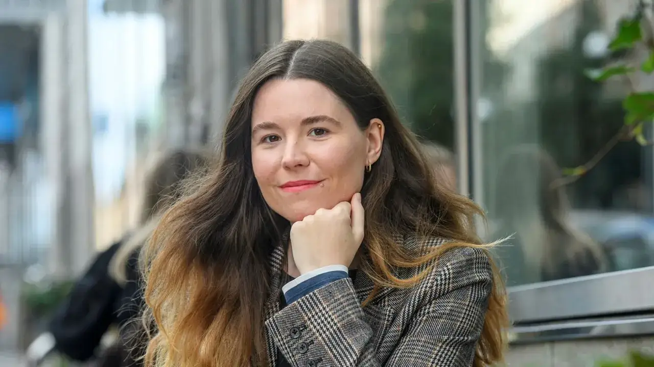 Johanna Hedman, med sitt långa, vågiga hår och en rutig kavaj, ler mot betraktaren.