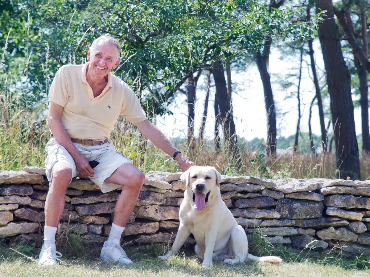 En leende man sitter på en stenmur med sin labrador. Kanske läser han ur sin jan eliasson bok i solen.