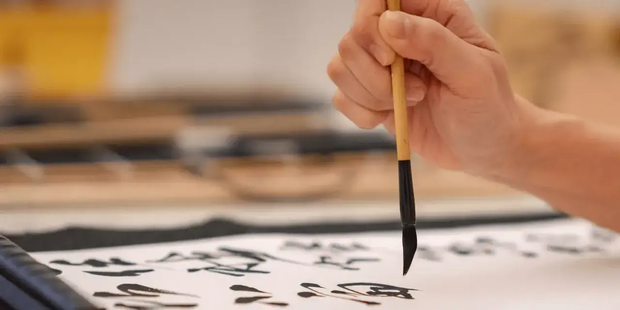 En hand h&aring;ller en pensel &ouml;ver ett papper med kalligrafi. Fri vers.