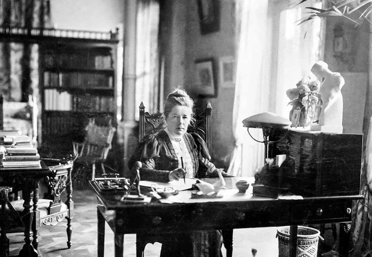 Selma Lagerlöf sitter vid sitt skrivbord, omgiven av böcker och skrivmaterial, redo att skriva sin nästa berättelse.