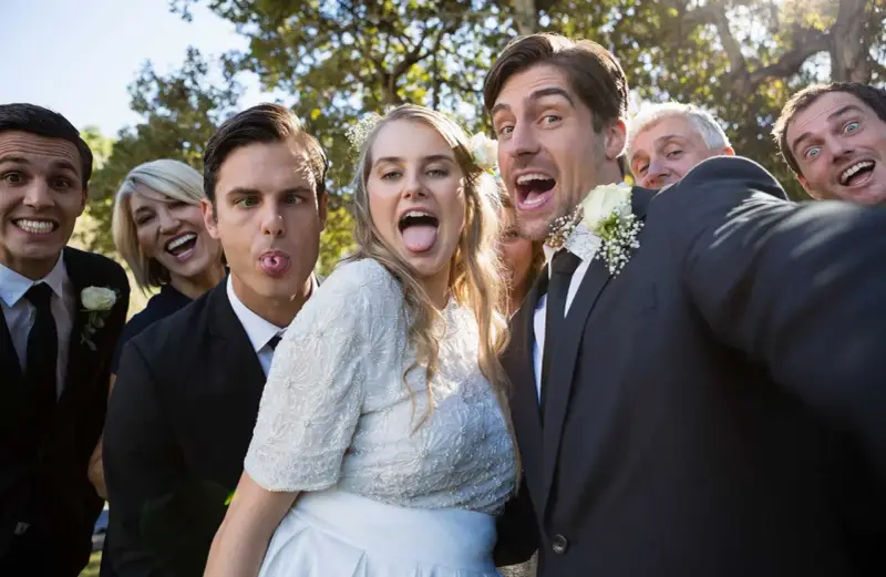 A wedding party takes a silly selfie outdoors, with the bride and groom sticking out their tongues.